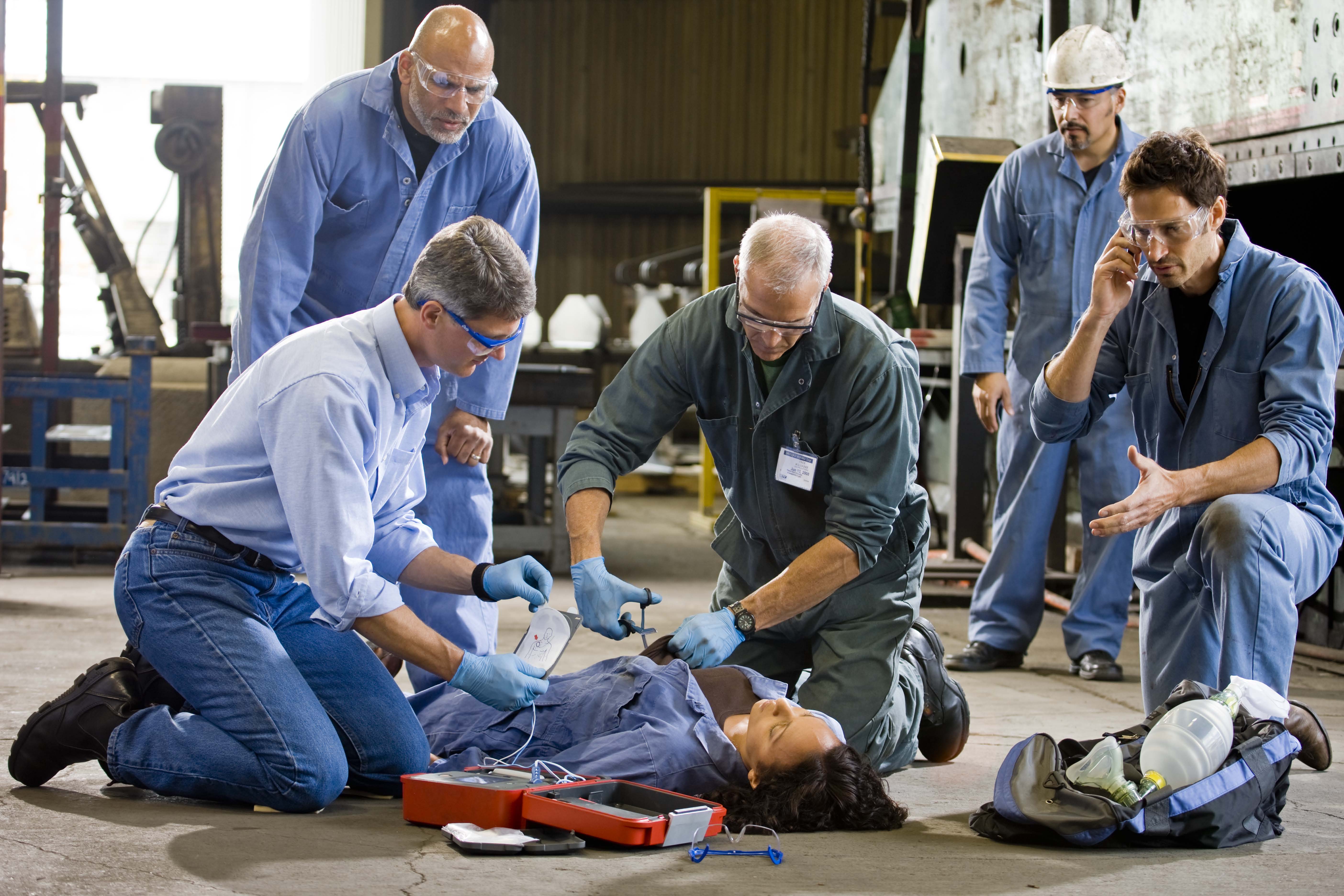 Workers in a factory using a defibrillator on a collapsed colleague while another calls for help