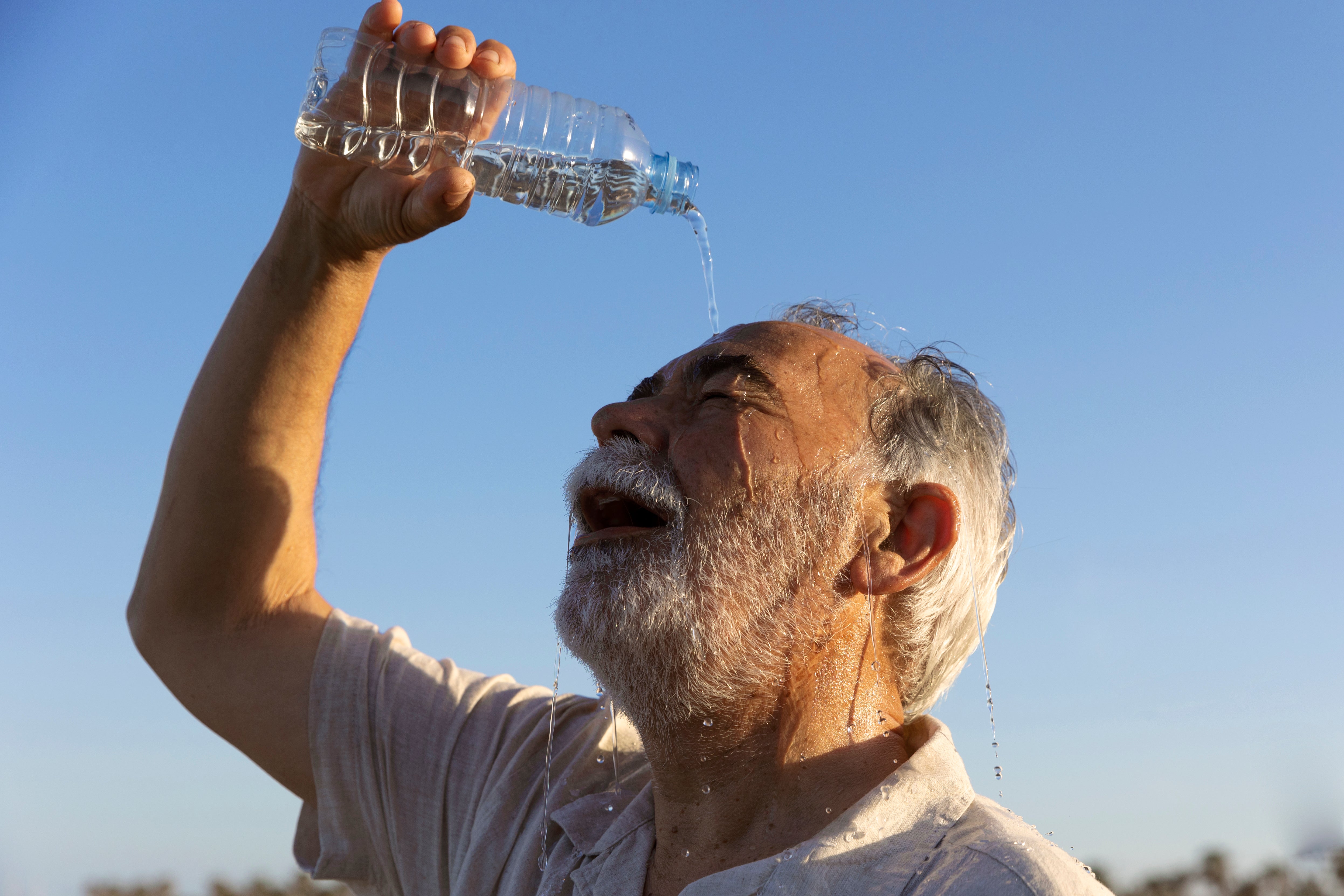 An older man pours water from a bottle over his face to cool down on a hot day under a clear blue sky.